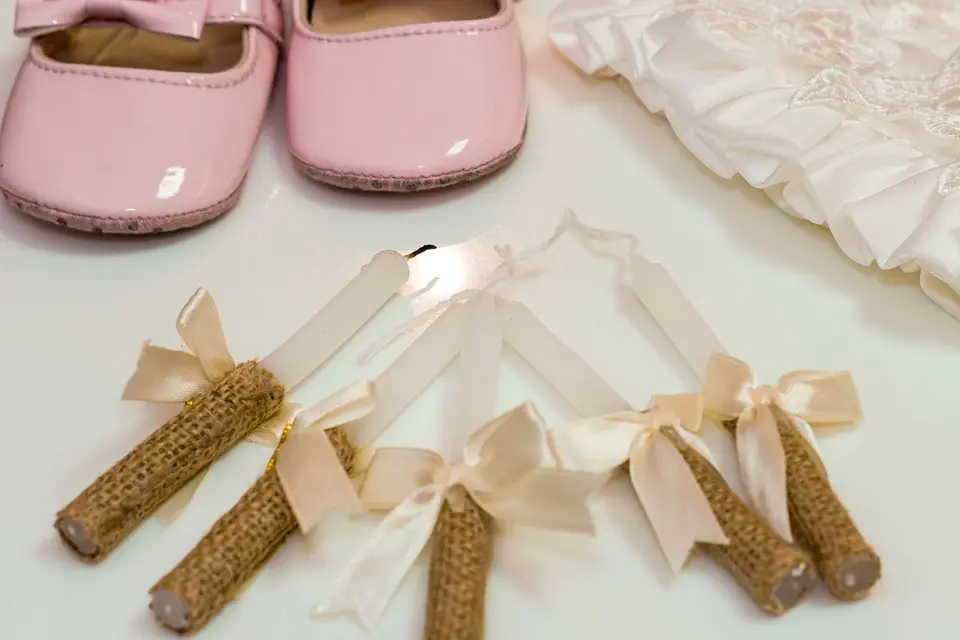 Zapatitos rosas de bebé con lazo, una prenda blanca bordada y velas con cintas, preparados para un bautizo