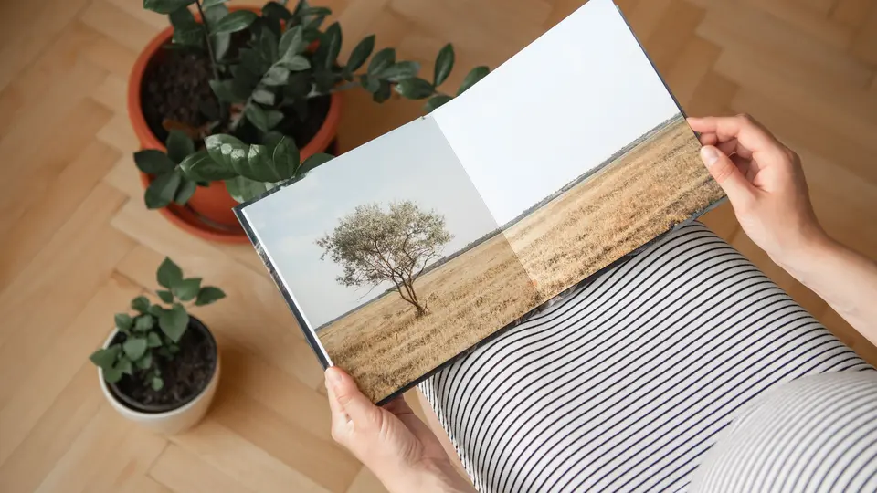Unas manos sostienen un álbum fotográfico abierto sobre las rodillas, con plantas y un suelo de madera al fondo