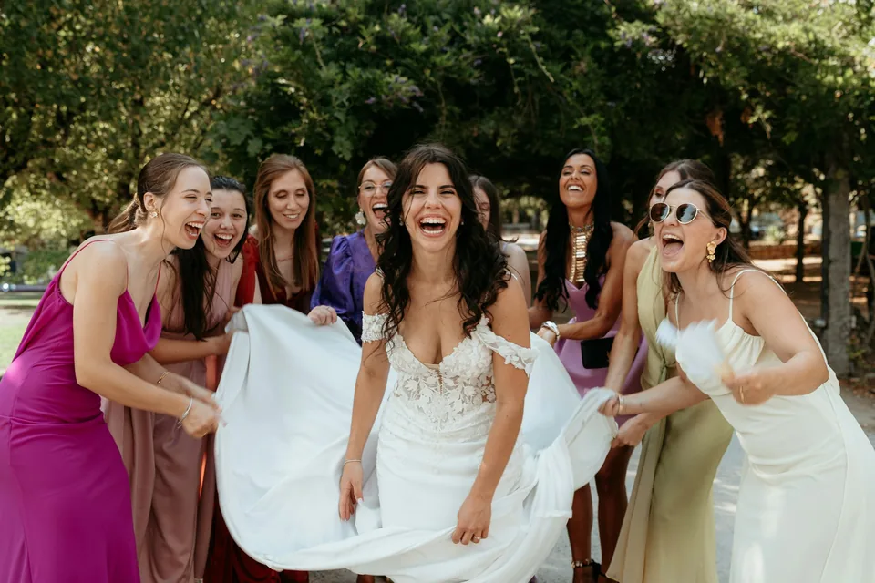 La novia ríe con sus amigas mientras le sujetan el vestido en el jardín