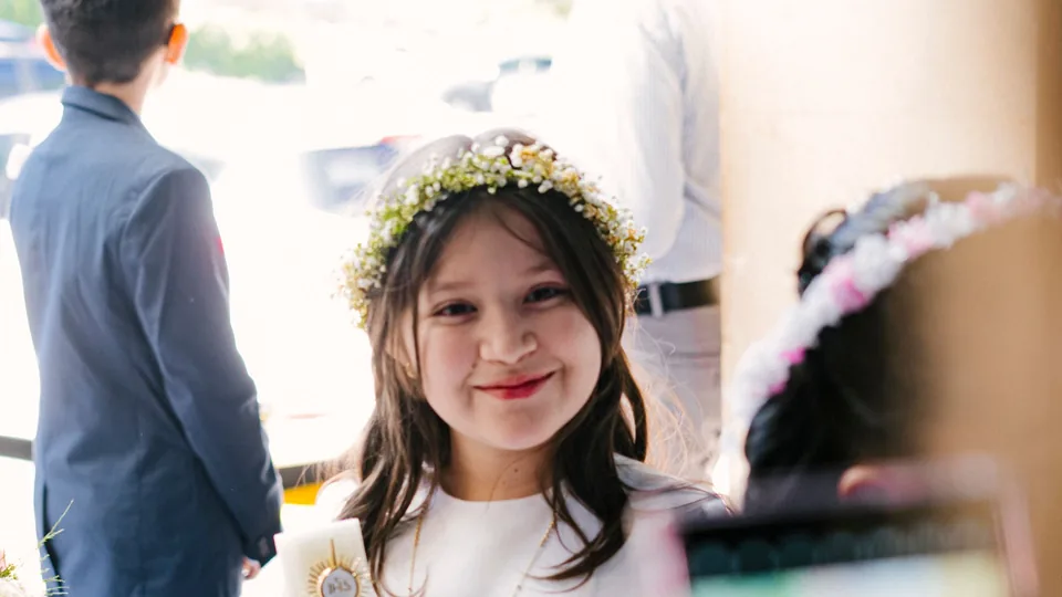 Una niña con corona de flores sonríe en su primera comunión mientras alguien la graba con el móvil