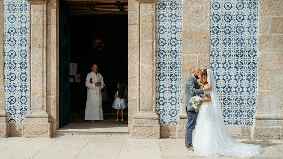 Los novios se abrazan a la puerta de una iglesia con azulejos mientras el sacerdote y una niña los miran
