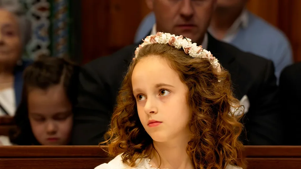 Una niña vestida de primera comunión sentada en el banco de la iglesia junto a su familia