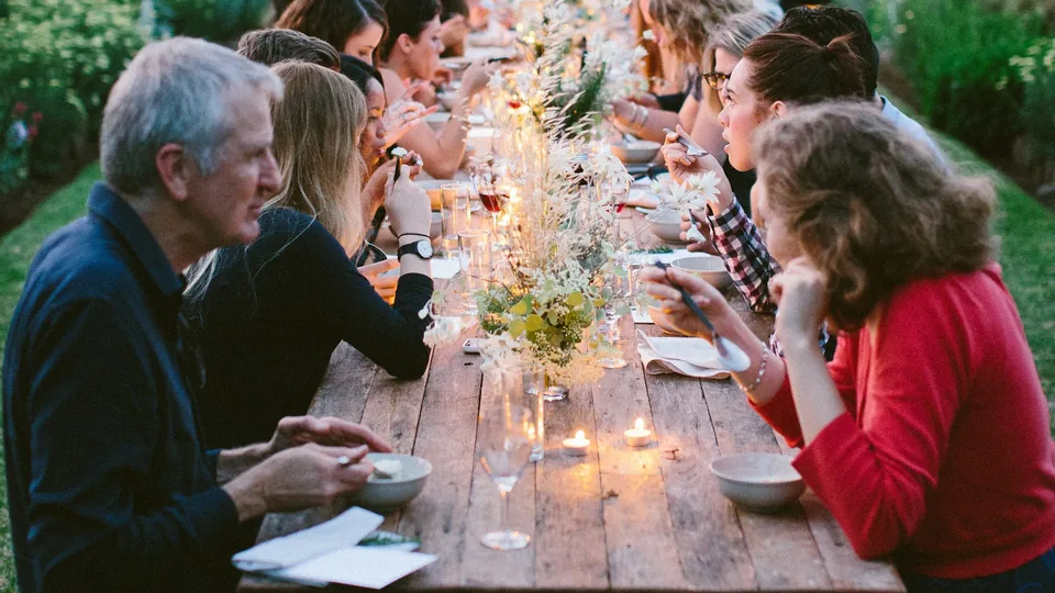 Invitados conversan en una larga mesa al aire libre durante una cena de celebración al atardecer