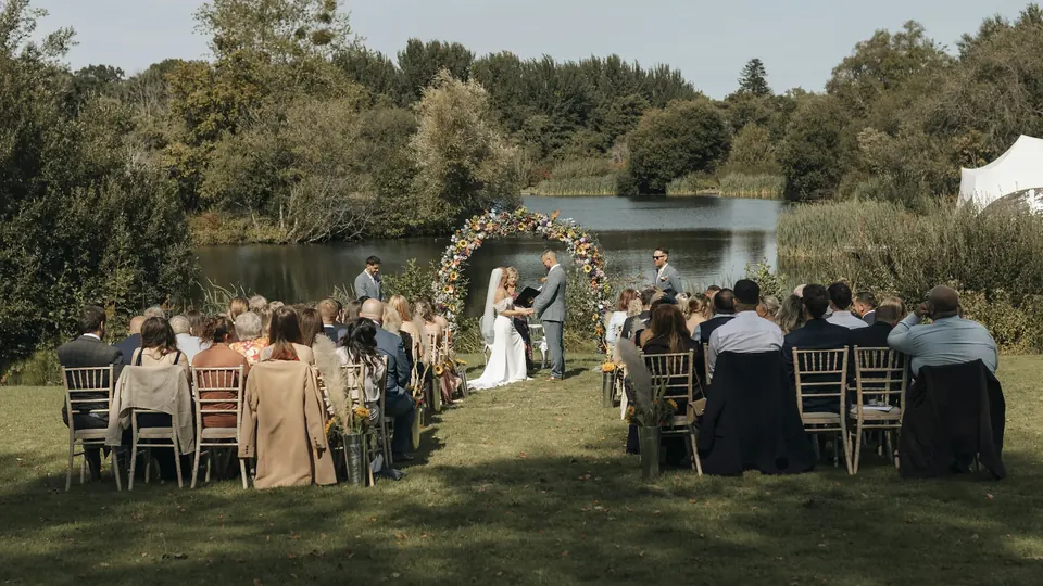 Ceremonia de boda al aire libre junto a un lago, con los invitados sentados ante el arco de flores