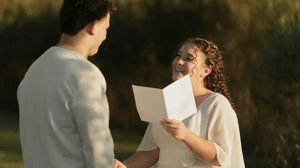 La novia lee sonriendo una carta escrita frente al novio al atardecer