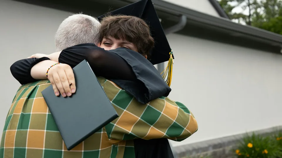 Un graduado con birrete y diploma abraza a un familiar tras la ceremonia