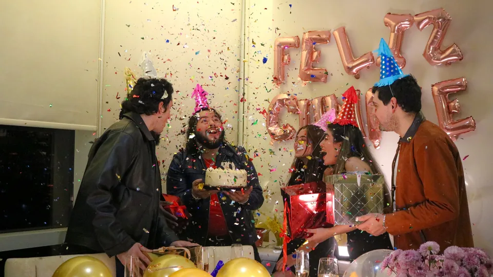 Un grupo de amigos lanza confeti alrededor de una tarta con velas ante unos globos que dicen «Feliz cumple»