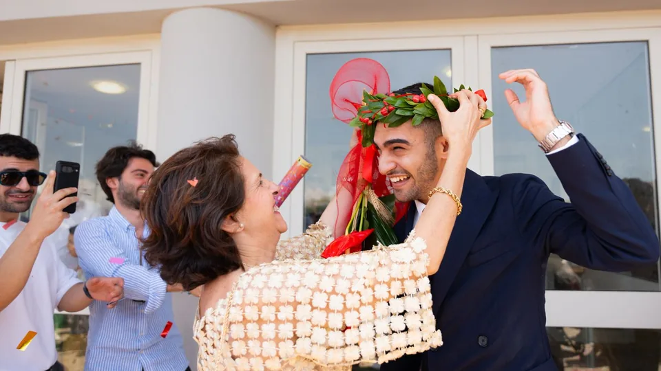 Una madre corona con laurel a su hijo recién graduado entre amigos y confeti a la salida de la universidad