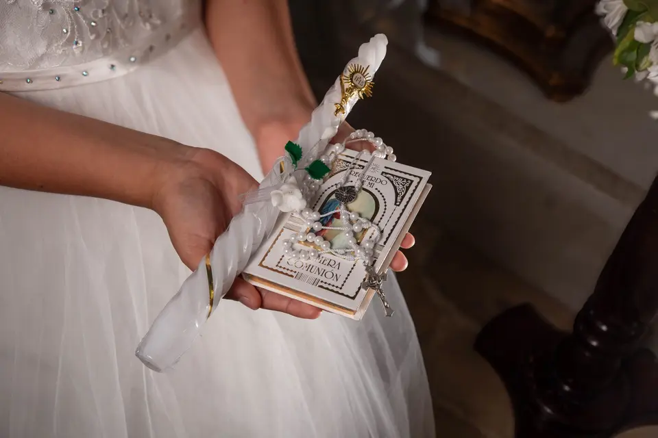 Unas manos sostienen una vela, un rosario y un librito de recuerdo de Primera Comunión sobre un vestido blanco