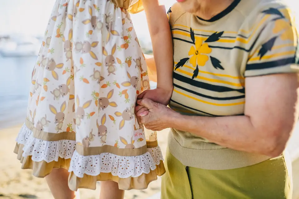 Una abuela y su nieta caminan de la mano en la playa; solo se ve la mano, la blusa y la falda
