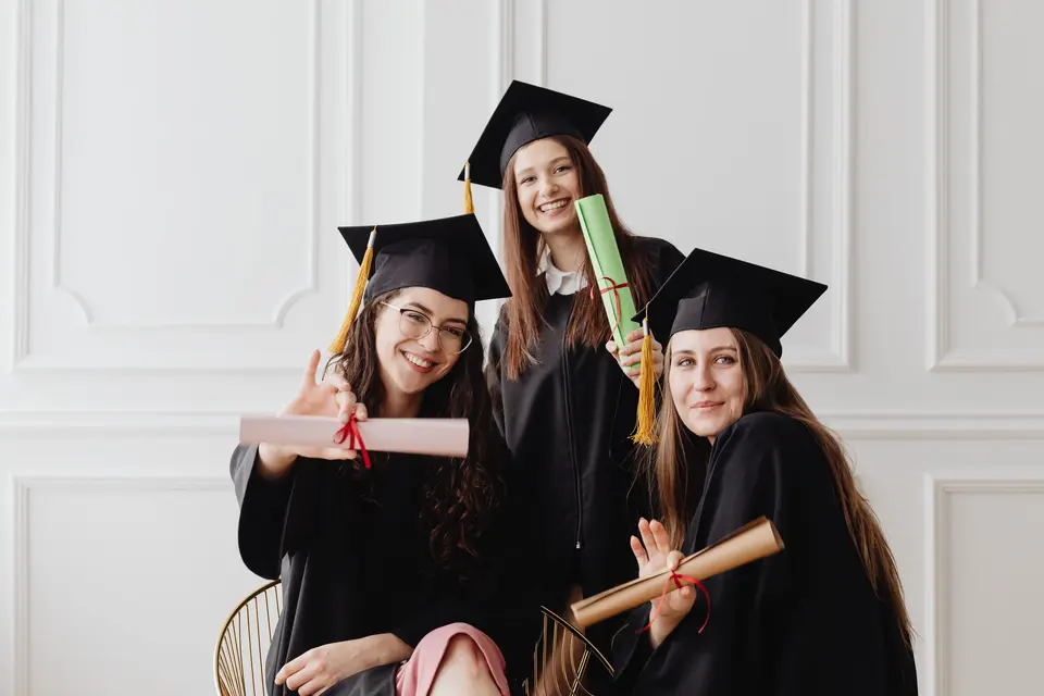 Tres amigas con toga y birrete posan abrazadas en una sala luminosa, con sus diplomas en las manos