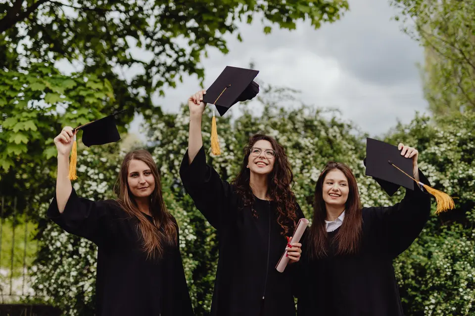 Tres amigas con toga lanzan los birretes al aire en un jardín, con sus diplomas en la mano