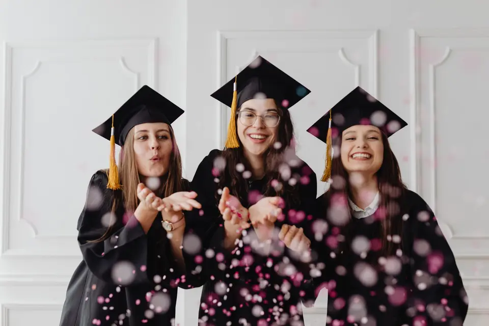Tres graduadas con toga y birrete soplan confeti rosa en una sala luminosa