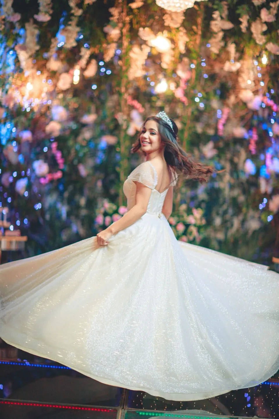 Una quinceañera con vestido blanco gira sonriendo en un jardín de flores y luces